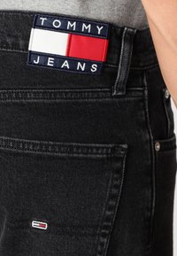 Jeans din denim negru cu un patch logo TOMMY JEANS, care prezintă elemente rectangulare roșii, albe și albastre; cusături detaliate și o buzunar din spate vizibil.