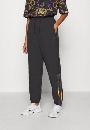 Personne portant un pantalon de survêtement noir avec des bandes arc-en-ciel et un logo sur la jambe, associé à des baskets blanches et une chemise à manches courtes à motifs.