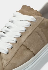 Μπεζ αθλητικό παπούτσι από σουέντ με λευκά κορδόνια, υφασμάτινο σχέδιο και κυματιστό περίγραμμα. Διαθέτει λευκή λαστιχένια εξωτερική σόλα και διακριτικό λογότυπο.