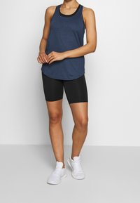 Námořnicky modrý sportovní tank top s kulatým spodním lemem přes černé cyklistické šortky. Modelka má na nohou bílé tenisky; látka působí měkce a prodyšně.