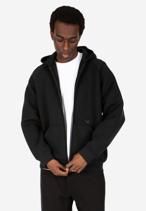 Man wearing black zip-up hoodie over white shirt, looking down while fastening zipper, standing against plain background.