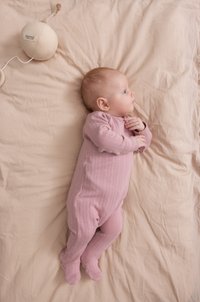 Un bébé en grenouillère rose repose sur une couverture crème, regardant sur le côté, à côté d'un jouet en peluche rond avec un cordon.