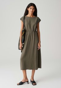 Olivgrünes, knielanges Kleid mit kurzen Ärmeln, geraffter Taille und strukturiertem Stoff. Kombiniert mit schwarzen Mules mit offenem Zehenbereich und einer geflochtenen Tasche.