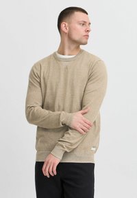 Beige gebreide trui met ronde hals en lange mouwen, met een gestructureerd patroon en een klein logo label aan de zoom. Draag met donkere broeken.