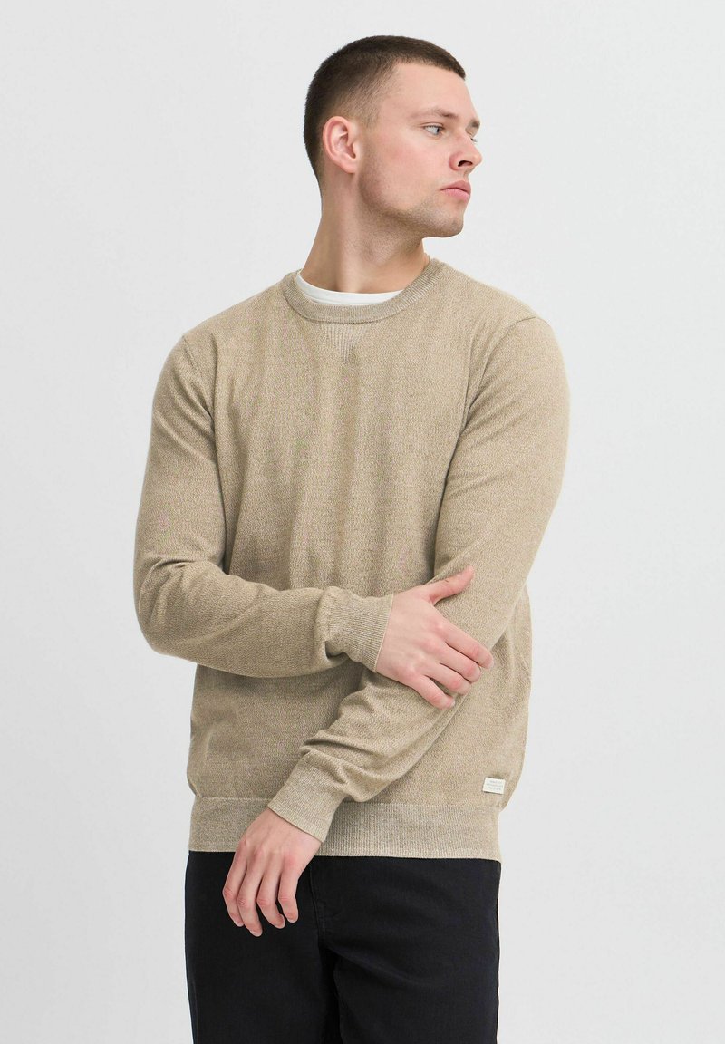Beige gebreide trui met ronde hals en lange mouwen, met een gestructureerd patroon en een klein logo label aan de zoom. Draag met donkere broeken.
