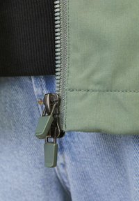 Zbliżenie podwójnego zamka błyskawicznego z zielonymi uchwytami na częściowo zapiętej zielonej kurtce, noszonej na jasnoniebieskich dżinsach.