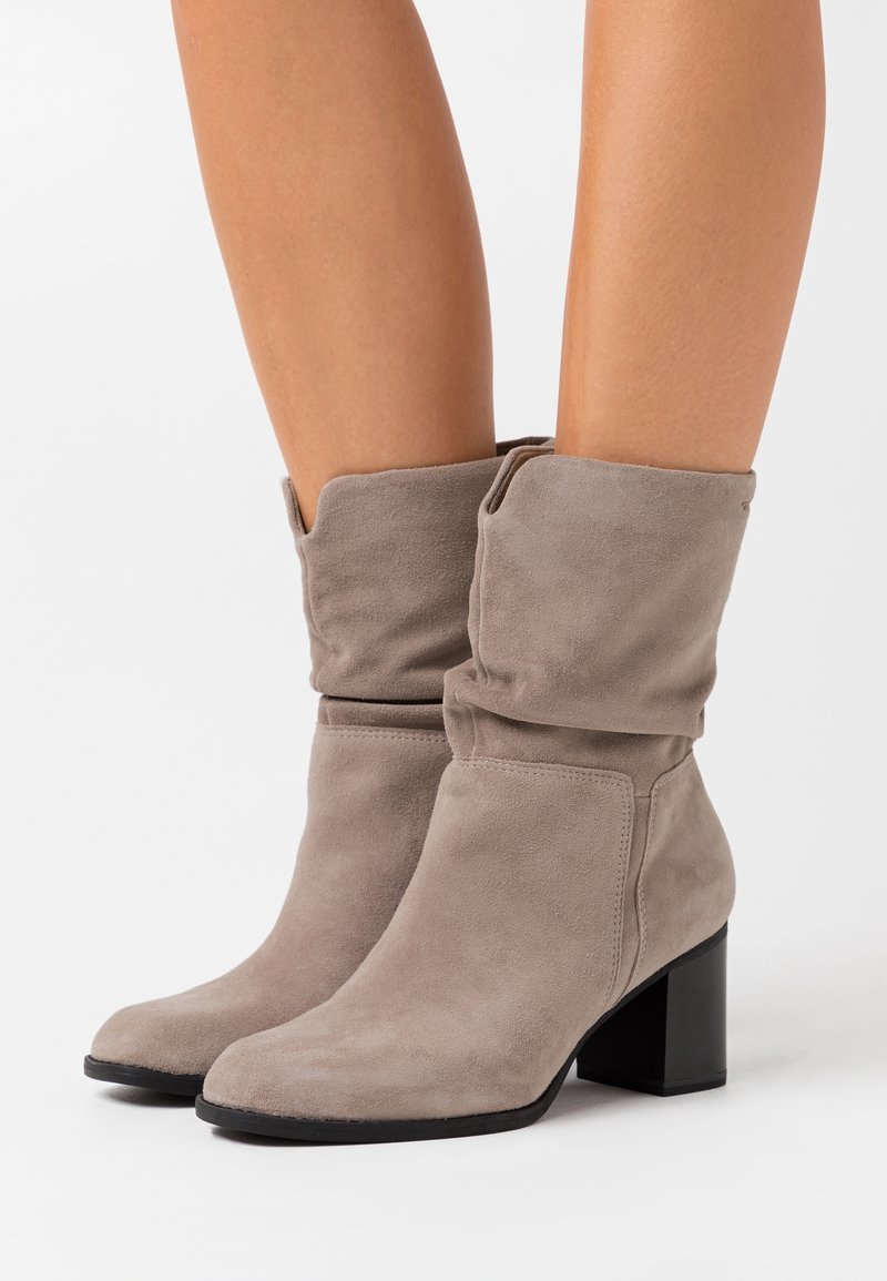 Paire de bottes mi-mollet en daim taupe à talons blocs portées sur jambes nues, avec un design souple et décontracté et des semelles noires.