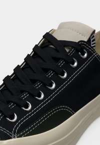 Μαύρα πάνινα παπούτσια με πράσινη λεπτομέρεια, λευκή ραφή και μεταλλικές τρύπες για τα κορδόνια· στρογγυλερή μύτη, επίπεδη σόλα και μπεζ λεπτομέρειες στη γλώσσα.