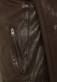 Giacca in pelle marrone con finitura lucida, cuciture visibili e tasca con zip. La texture è liscia e il design è aderente.