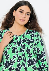 Zielona bluzka z czarnym kwiatowym wzorem, okrągłym dekoltem i teksturowanym materiałem. Projekt zawiera subtelne fałdy i krótkie rękawy.