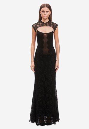 Woman wearing a floor-length black lace gown with a sheer front panel and cap sleeves, standing against a white background.