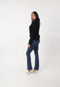 Suéter negro de punto con mangas largas, combinado con jeans oscuros de corte acampanado. El modelo lleva zapatos de tacón beige, mostrando un perfil lateral.