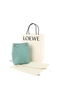 Sac en cuir Loewe vert clair avec boutons argentés, placé à côté d'un sac shopping blanc Loewe et de deux housses anti-poussière beige sur fond blanc.