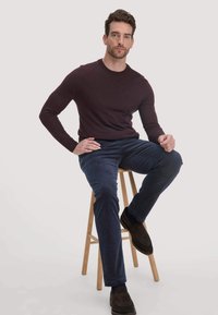 Rundhals-Maronpullover mit enganliegenden Ärmeln; kombiniert mit strukturierten navy-blauen Cordhosen und dunkelbraunen Slippern, sitzt auf einem Holzschemel.