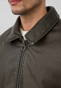 Braune Lederjacke mit einem Kragen, ausgestattet mit einem schwarzen Reißverschluss und einer glatten Textur, gezeigt von der Schulter bis zur Kinnhöhe.