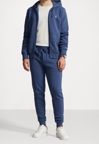 Blå bomullshoodie med dragkedja, två framfickor, matchande blå sweatshirtbyxor och vita sneakers. Slim fit med dragsko i midjan.