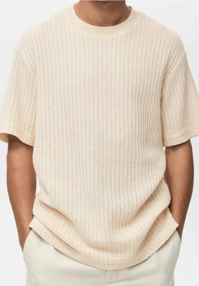 Chemise beige à manches courtes en maille texturée portée par une personne avec les mains dans les poches d'un pantalon de couleur claire.
