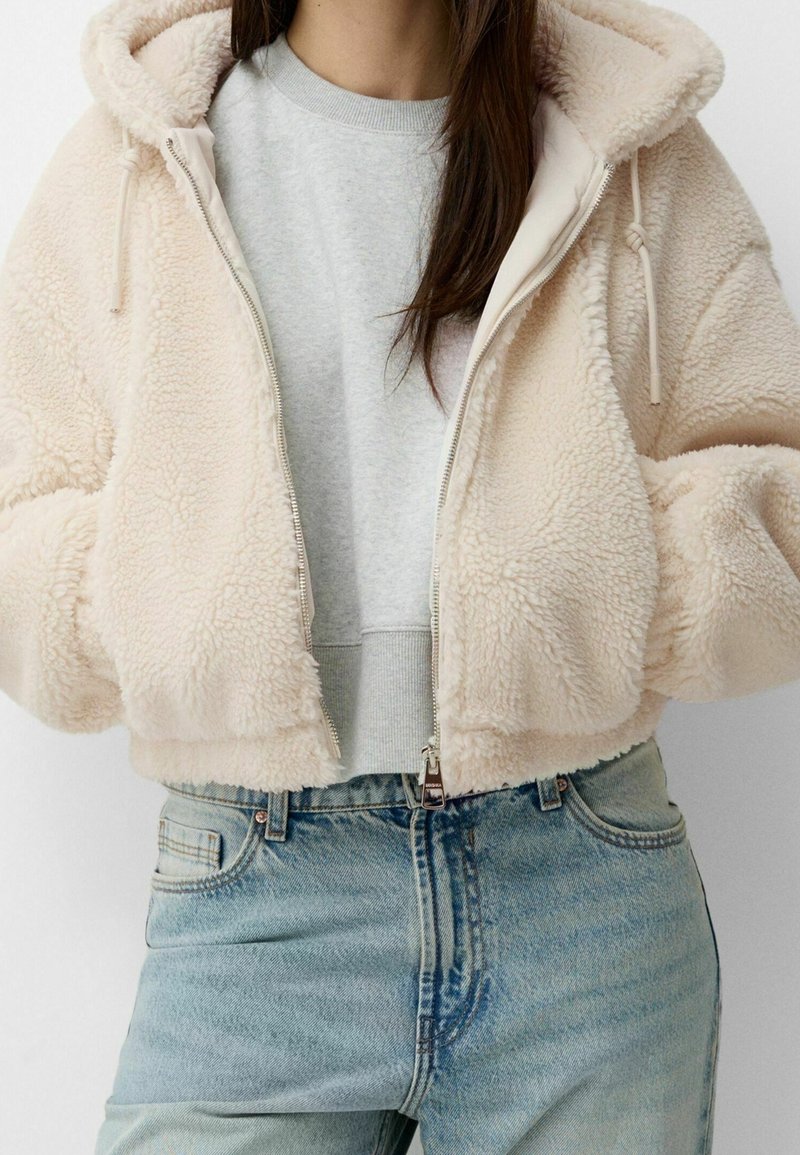 Personne portant un sweat à capuche crème, duveteux et zippé, sur un sweat-shirt gris clair et un jean denim bleu clair, les mains dans les poches de la veste.