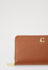 Portefeuille en cuir marron avec une surface texturée, accents en métal doré et un logo distinctif. Il possède une fermeture éclair et une forme rectangulaire.