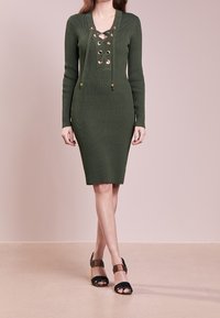 Donna che indossa un abito in maglia a coste verde oliva con maniche lunghe e dettagli con lacci sul davanti, abbinato a sandali aperti neri e marroni.