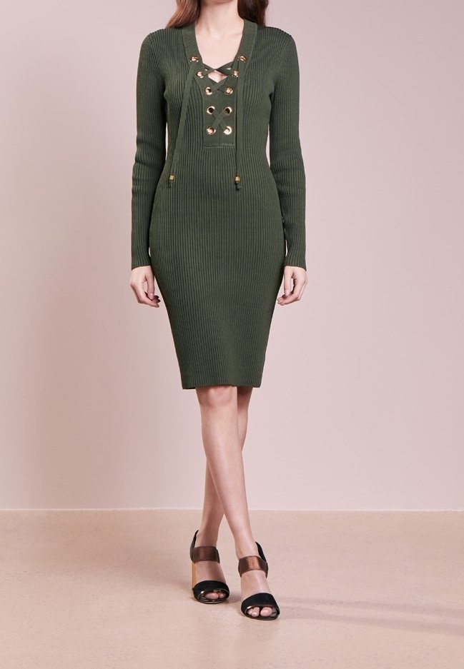 Donna che indossa un abito in maglia a coste verde oliva con maniche lunghe e dettagli con lacci sul davanti, abbinato a sandali aperti neri e marroni.