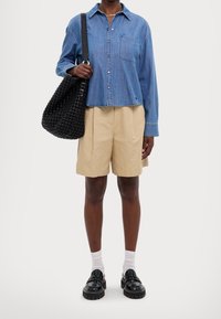 Ljusblå denimskjorta med knappar och bröstficka, beige plisserade shorts, svart vävd handväska och glänsande svarta skor med tjocka sulor.