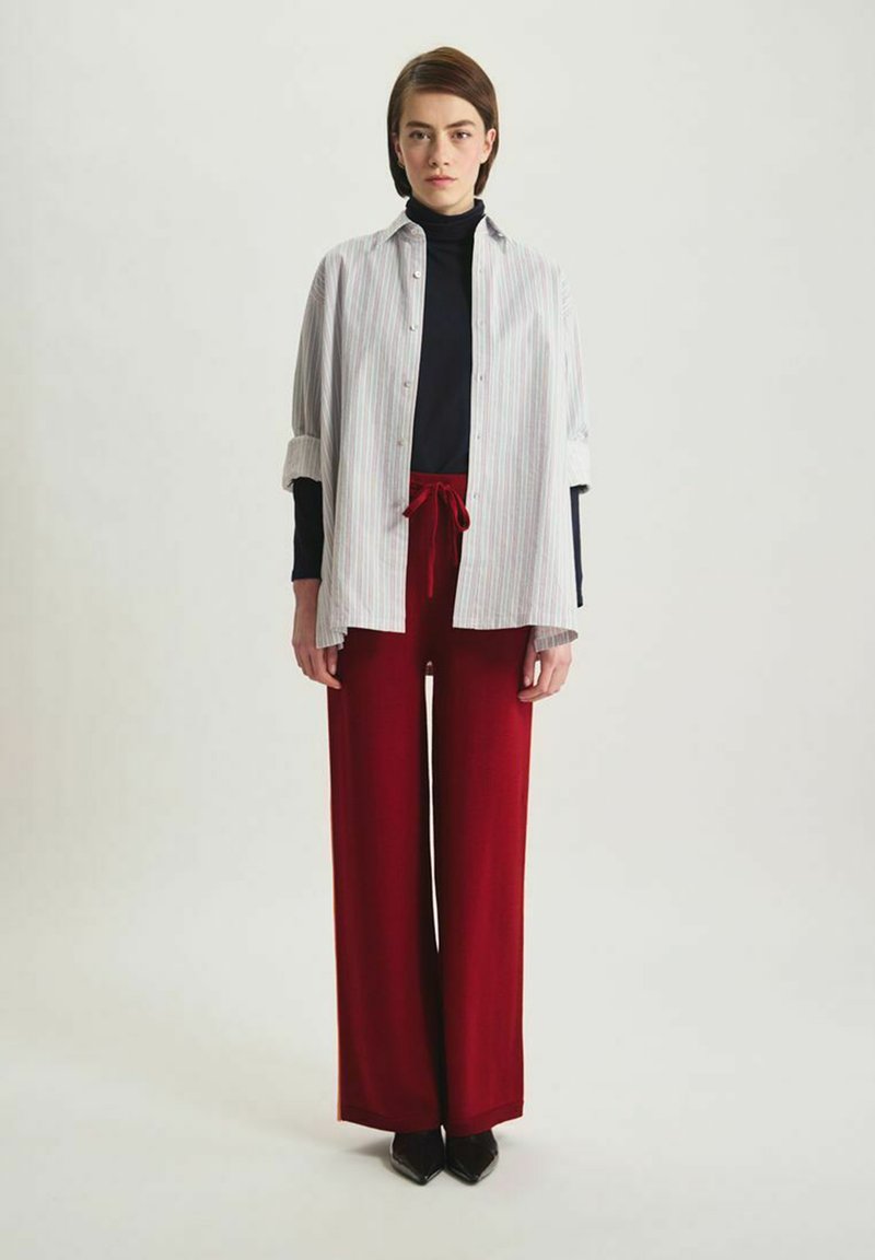 Chemise blanche et rayée à boutons superposée sur un col roulé noir, associée à un pantalon large rouge taille haute et des chaussures noires à bouts pointus.