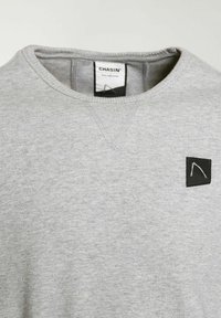 Sudadera gris con cuello redondo, que presenta una etiqueta de tela negra en el pecho y un tejido de punto texturizado.