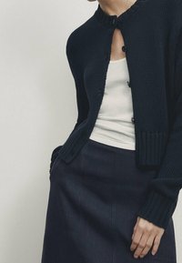 Marineblauwe gebreide cardigan met een ronde hals, knoopsluiting en geribbelde accenten. Gedragen over een wit hemdje en gecombineerd met een donkere rok.