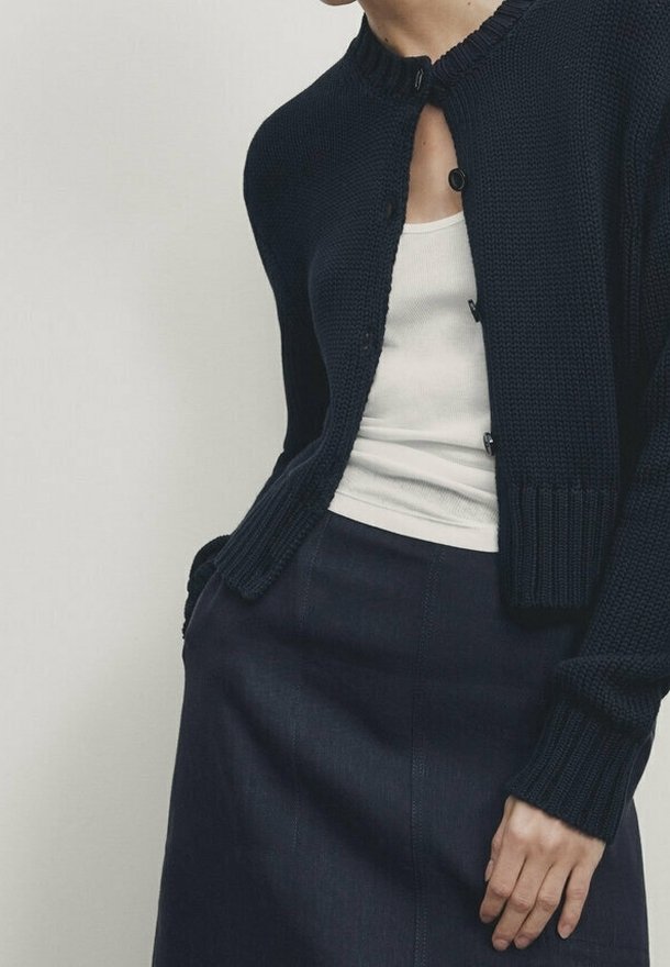 Marineblauwe gebreide cardigan met een ronde hals, knoopsluiting en geribbelde accenten. Gedragen over een wit hemdje en gecombineerd met een donkere rok.