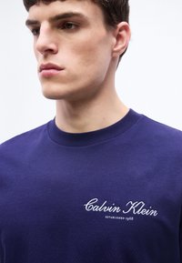 Νεαρός άνδρας που φοράει μπλε ναυτικό πουκάμισο με λευκή κέντηση "Calvin Klein Established 1968" στο στήθος, κοιτάει ελαφρώς προς τα αριστερά.