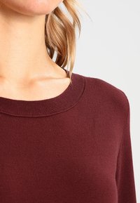 Pull bordeaux avec un col rond, une texture douce et des bordures côtelées. Le matériau semble léger et lisse, parfait pour superposer.