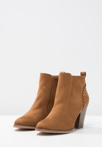 Botines de ante marrón con textura suave, puntera redondeada y tacón apilado. Incluye detalles de costura lateral y una lengüeta en la parte posterior.