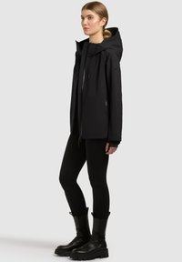 Giovane donna in piedi, che indossa una giacca nera con cappuccio, leggings neri e stivali neri con suola alta, su uno sfondo chiaro e uniforme.