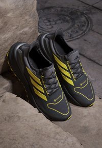 Scarpe da corsa grigie con accenti gialli, materiale GORE-TEX e pannelli testurizzati. Presentano una suola spessa e un design a tre strisce.
