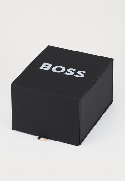 Boîte rectangulaire noire avec une texture mate, portant le mot "BOSS" en lettres majuscules et en couleur argentée sur le couvercle. Petite étiquette de couleurs distinctes.