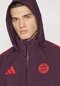 Giacca leggera viola con cappuccio, caratterizzata dal logo rosso Adidas e dall'emblema del FC Bayern Monaco. Chiusura con zip e cordino regolabile.
