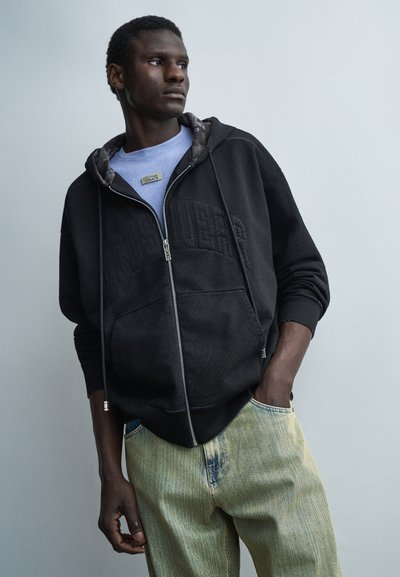 Joven con sudadera negra con cremallera sobre una camiseta azul y jeans de color claro, de pie con una mano en el bolsillo, mirando de lado.