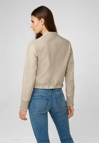 Beige Leder-Bomberjacke mit gerippten Bündchen und Saum, ausgestattet mit einer glatten Textur und einem strukturierten Kragen. Getragen mit blauen Jeans.