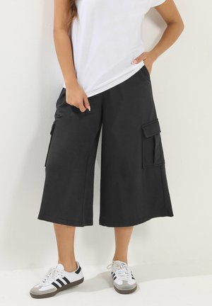 Donna che indossa pantaloncini cargo neri a gamba larga, t-shirt bianca e scarpe da ginnastica bianche con strisce nere, in piedi contro un muro bianco.