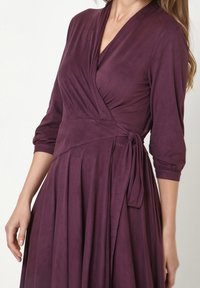 Violettes Wickelkleid mit Dreiviertelärmeln, aus weichem Stoff, mit plissiertem Rock und seitlichem Bindeband für eine verstellbare Passform.