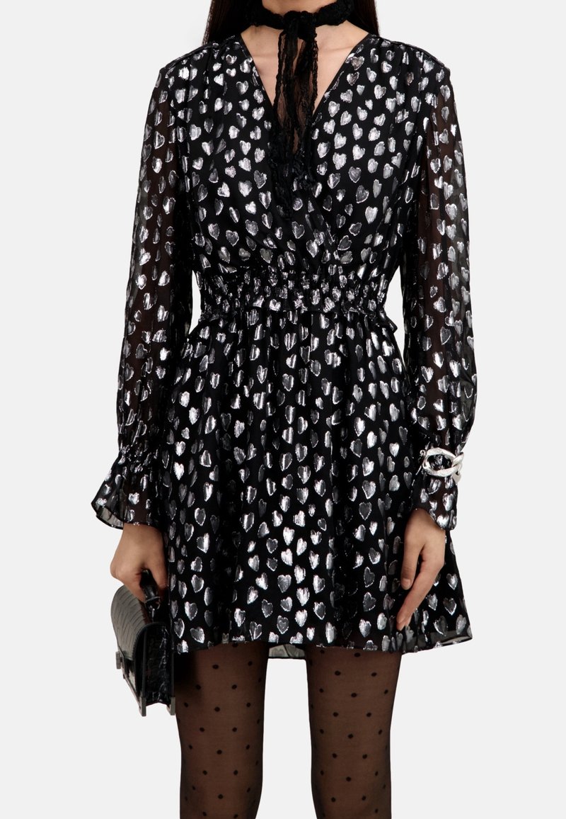 Robe noire à manches longues ornée de motifs cœurs argentés, manches transparentes, taille élastique, accompagnée de collants noirs à pois et d'une pochette noire.