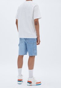 Camiseta de algodón blanca, pantalones cortos de mezclilla azul claro con bolsillos laterales, zapatillas blancas con acentos en naranja y azul, calcetines grises, visto desde la parte de atrás.
