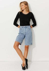Haut noir à manches longues, bermudas en denim bleu clair, et ballerines noires avec des clous en argent. Le modèle se tient devant un fond blanc.