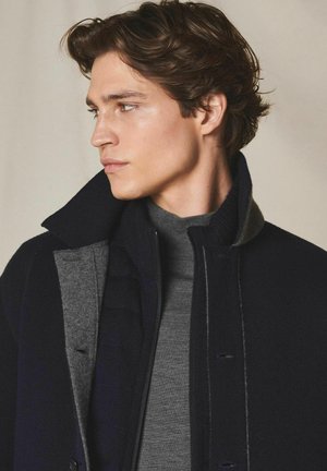 Junger Mann mit welligem braunem Haar, der einen schwarzen und grauen Mantel über einem grauen Rollkragenpullover trägt und nach links schaut, vor einem neutralen Hintergrund.