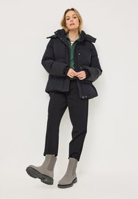 Veste en duvet noire avec capuche amovible, portée sur un haut vert, associée à un pantalon noir et des bottines grises à semelles épaisses.