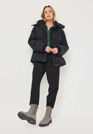 Svart pufferjakke med avtagbar hette, brukt over en grønn topp, sammen med svarte bukser og grå ankelboots med tykke såler.