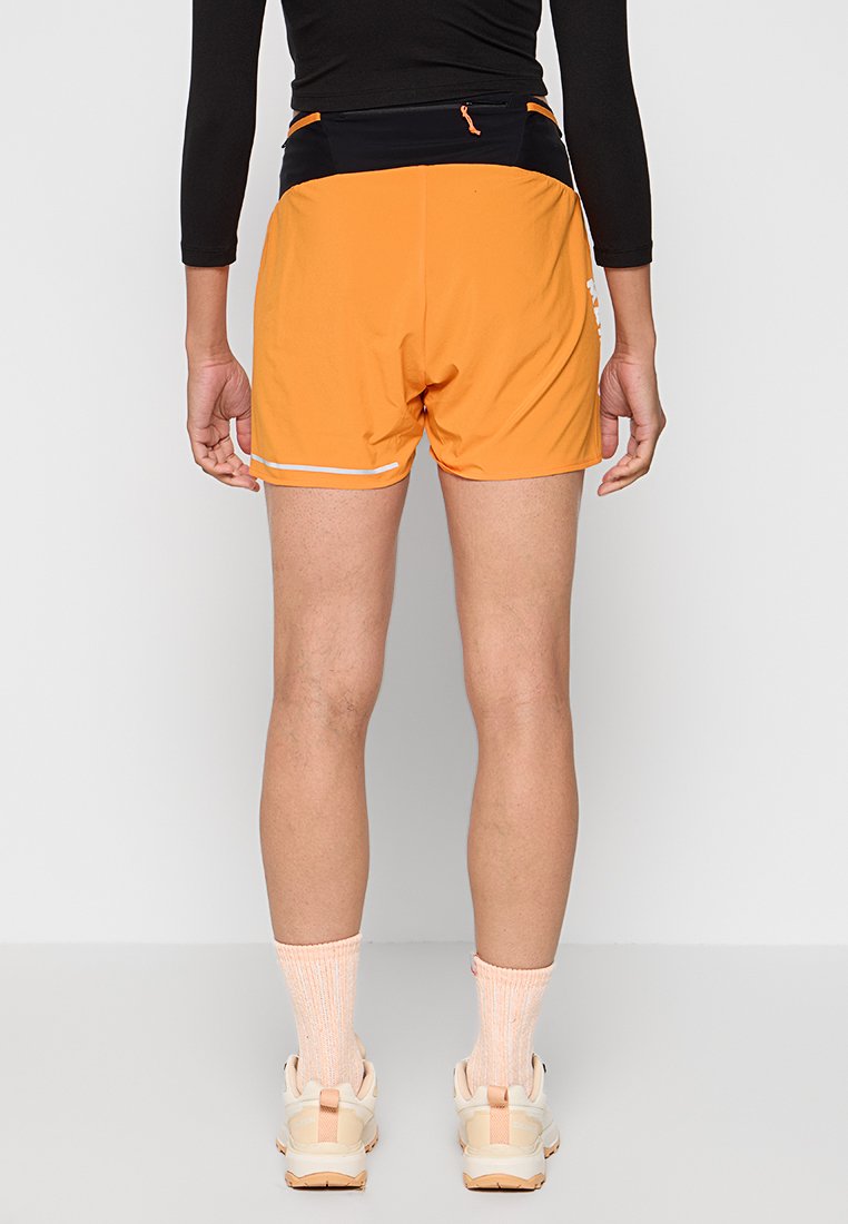 Pantalones cortos deportivos naranjas con una cinturilla negra, que cuentan con un bolsillo con cremallera y un ribete reflectante blanco a lo largo del dobladillo. Textura lisa.