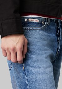 Jeans in denim azzurro chiaro con sfumature visibili, etichetta con la scritta "Calvin Klein", abbinati a una giacca nera, mano appoggiata sulla tasca.