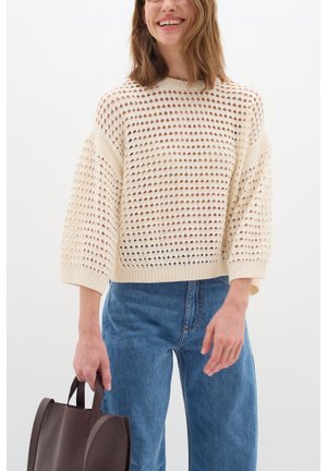 Strickpullover in Creme mit einem lockeren, durchbrochenen Muster, breite Ärmel und gerippte Kanten. Kombiniert mit hochtaillierten blauen Jeans und einer braunen Tasche.